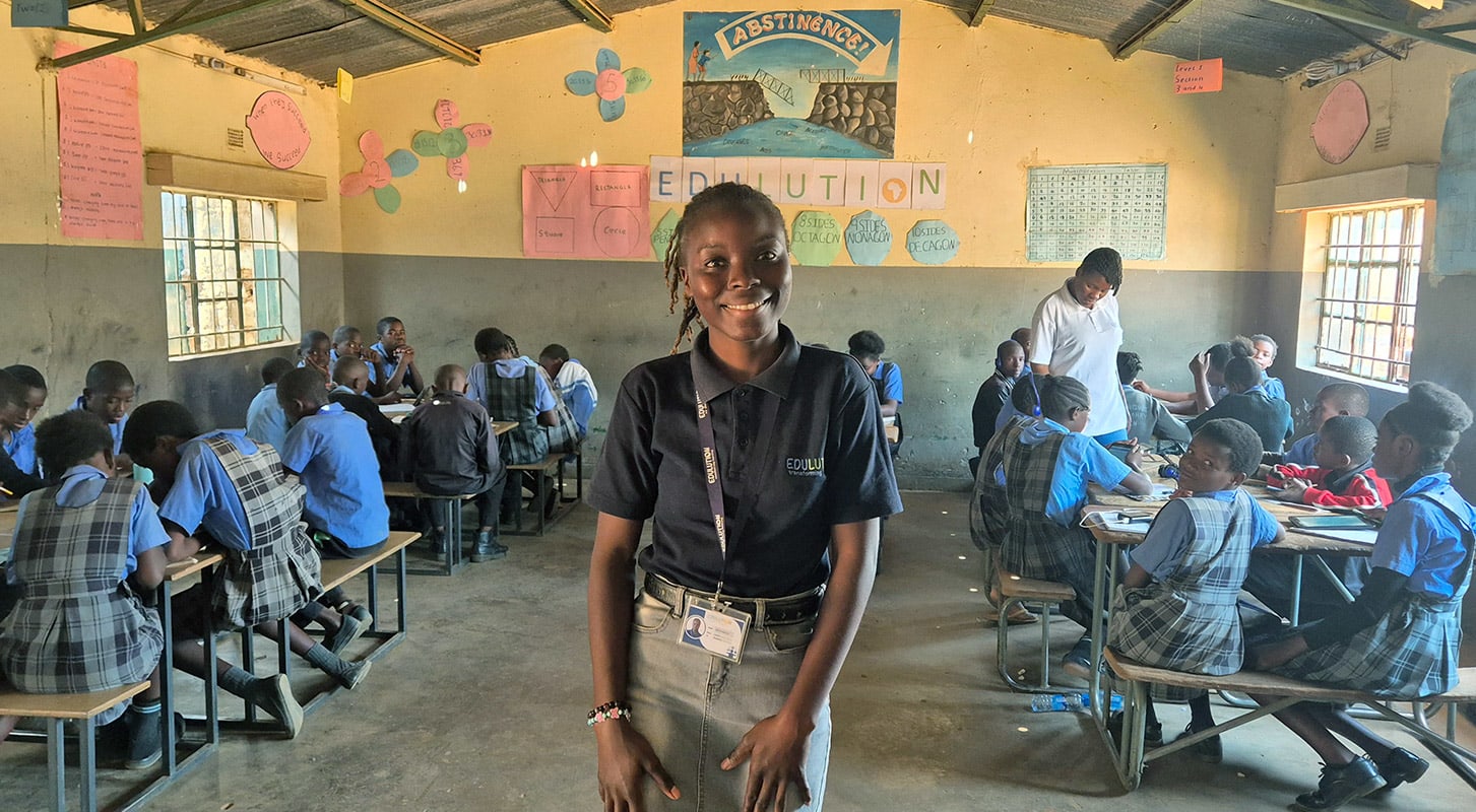 A girl standing in a classroom setting surrounded by children dressed in uniform, seated in rows of desks, participating in class activities.